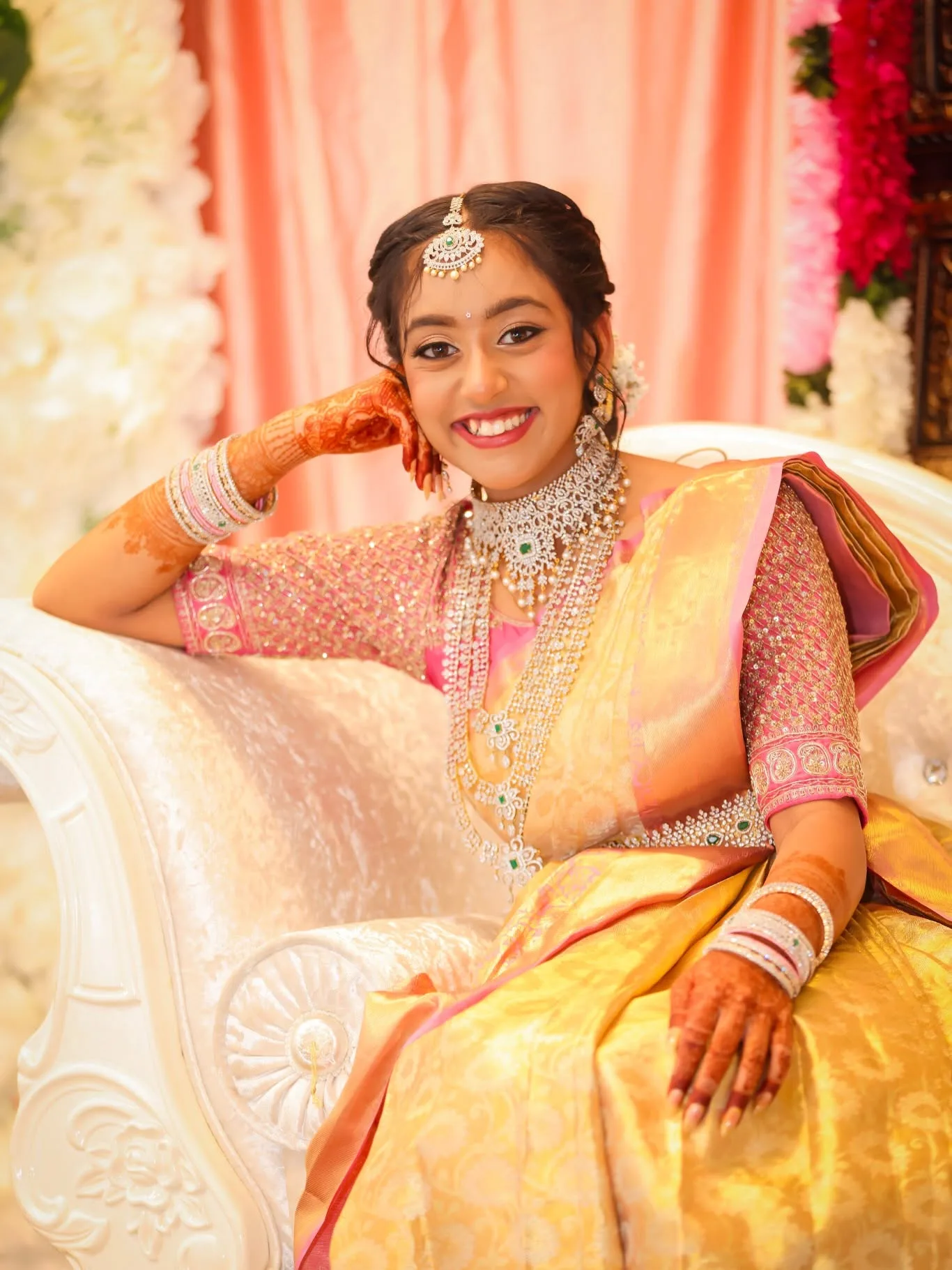 Bride seated on white settee in gold kanjivaram and diamond bridal jewellery, mehndi hands, floral backdrop