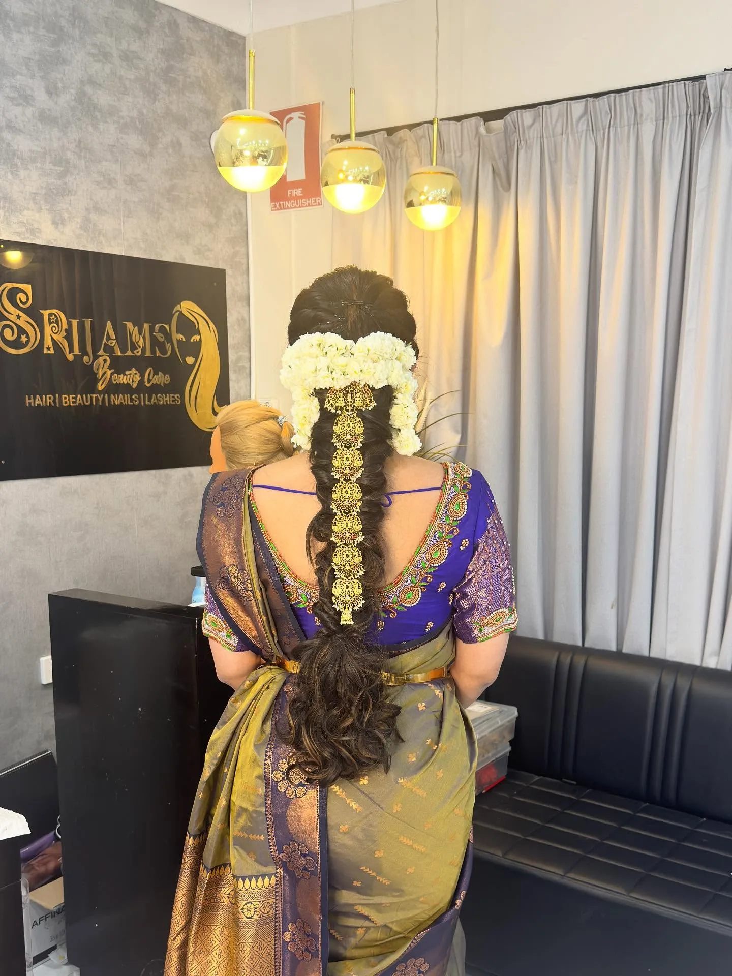 Rear view of bridal single-braid dressed with jasmine and gold jada, purple and olive saree, in front of the Srijams plaque