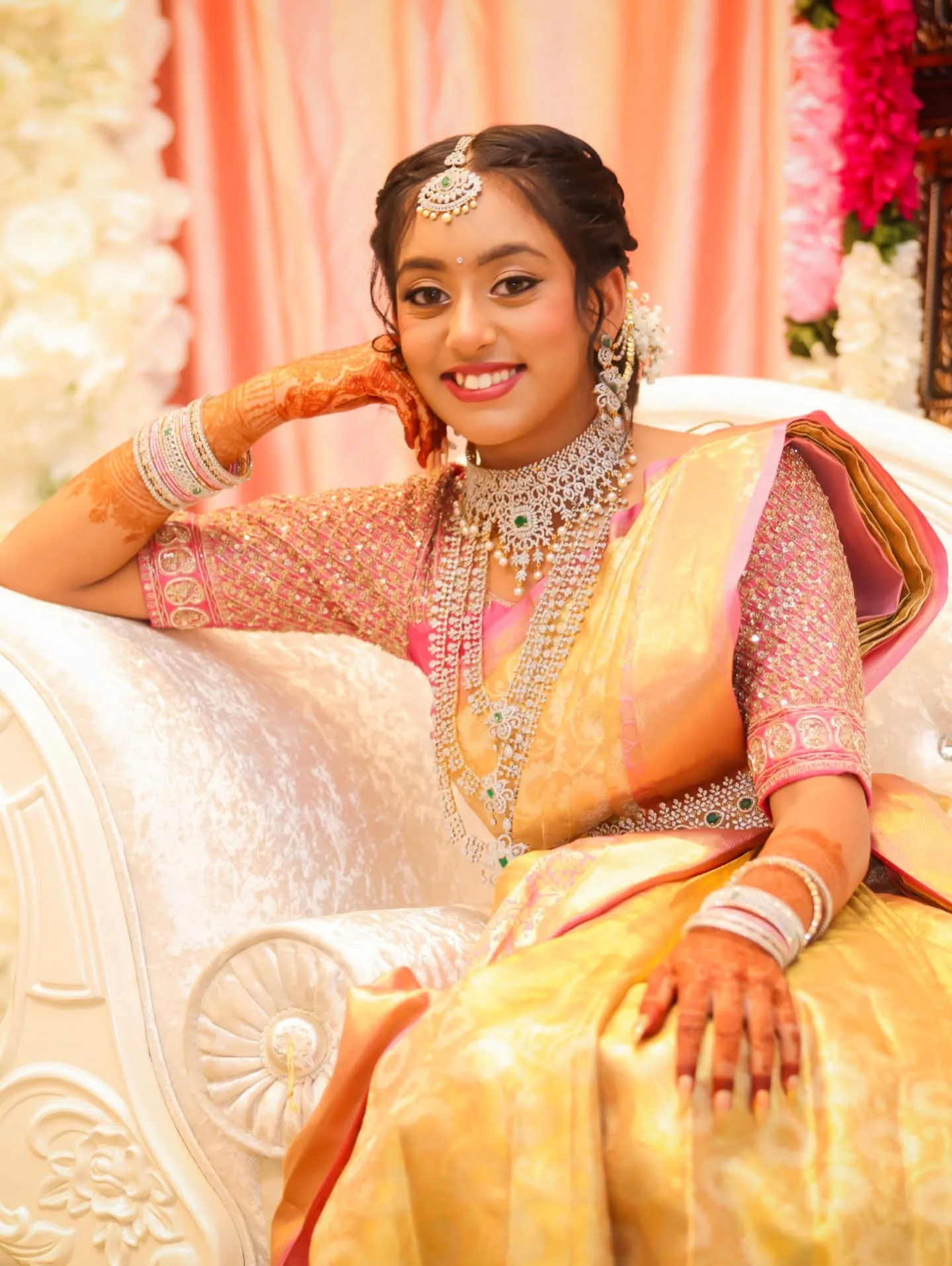 Bride seated in gold saree with layered diamond sets, mehndi, white floral backdrop
