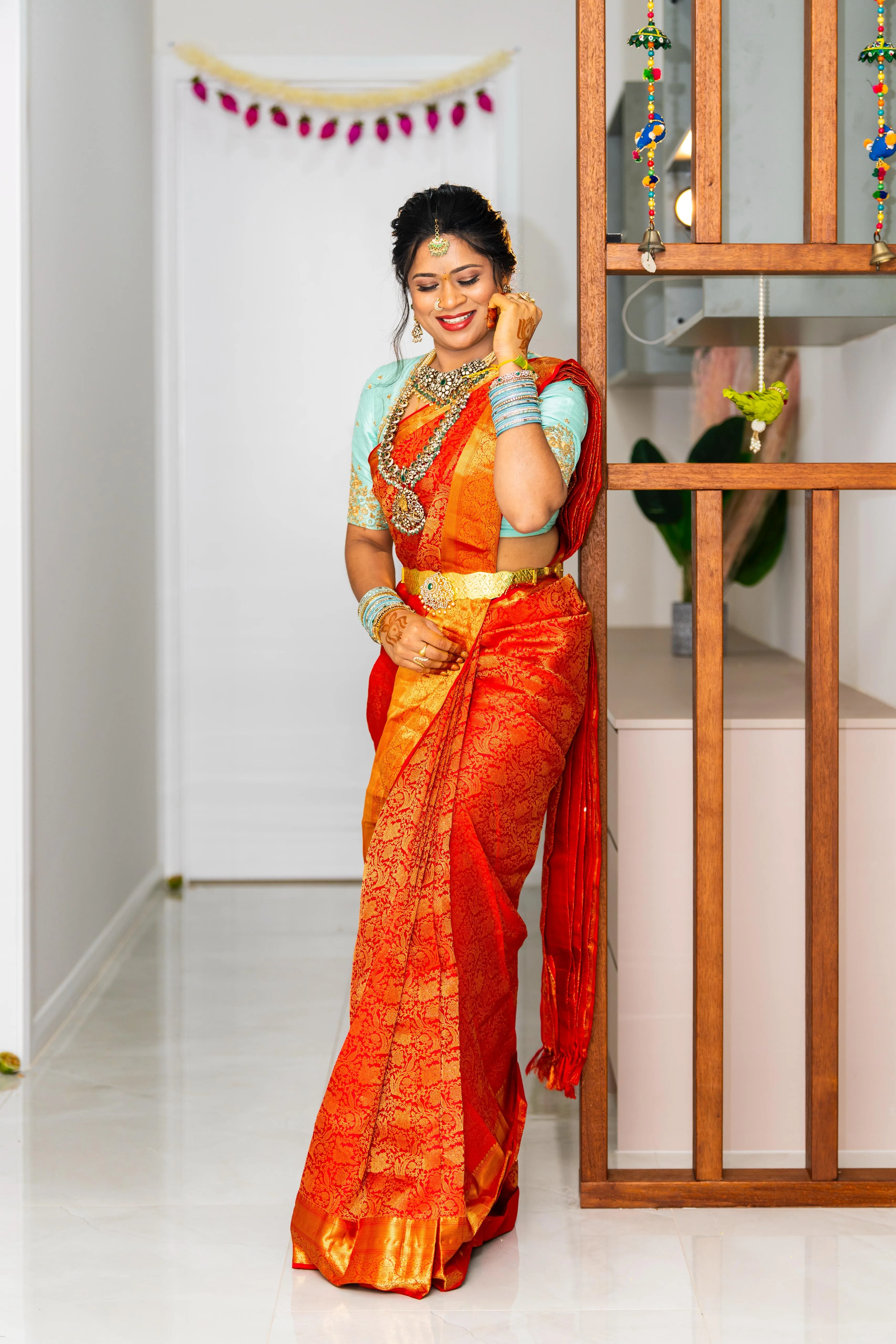 Full-length bridal portrait in orange kanjivaram and teal blouse, leaning on a timber screen at a festive home venue