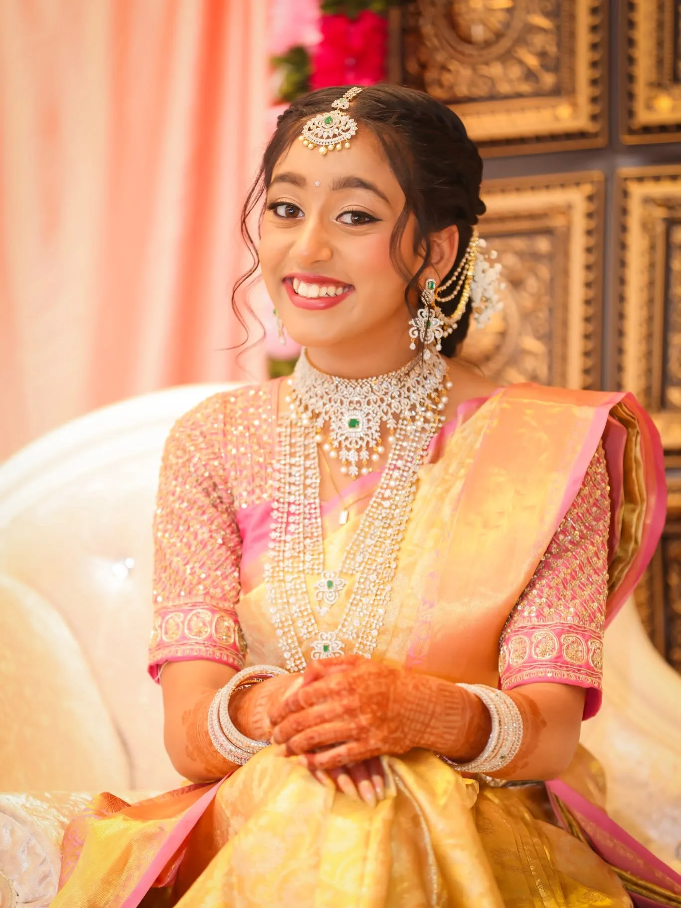Bride seated in pink and gold saree with diamond jewellery, warm gold backdrop
