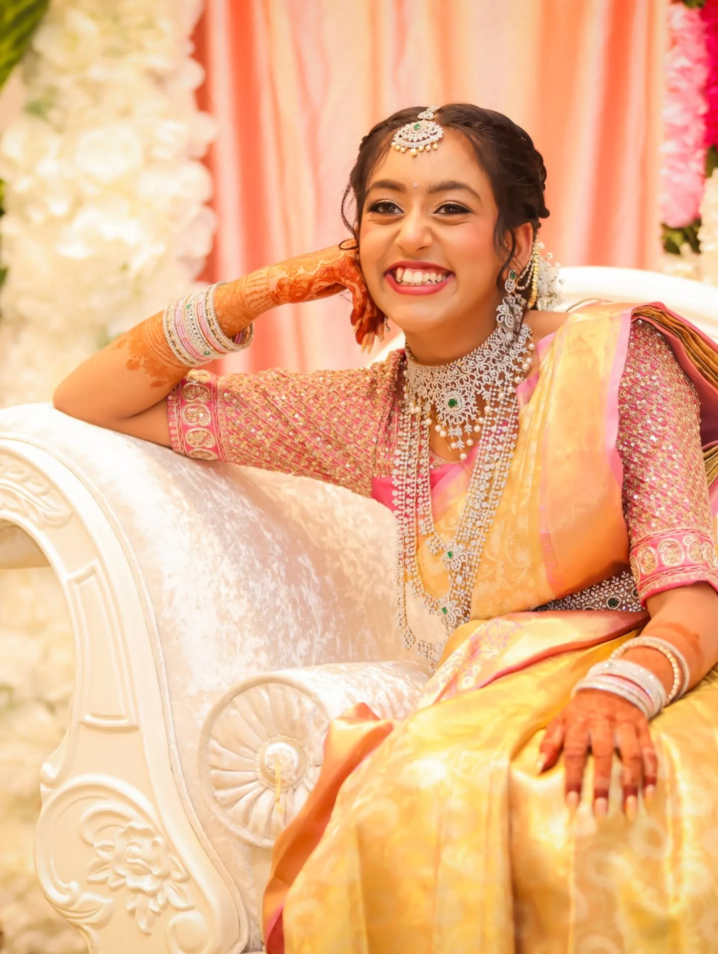 Bride seated in gold saree and layered diamond haaram, mehndi, white floral backdrop