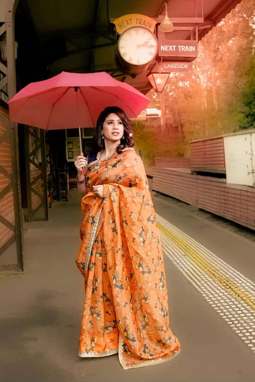 Styled client in orange floral saree with a red umbrella on a heritage train station platform